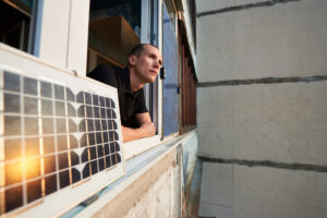 Junger Mann auf dem Balkon eines Wohnhauses mit Solarpanel, in dem sich die Sonne bei Sonnenuntergang spiegelt. Konzept zur Umwandlung von Sonnenstrahlung in Strom.