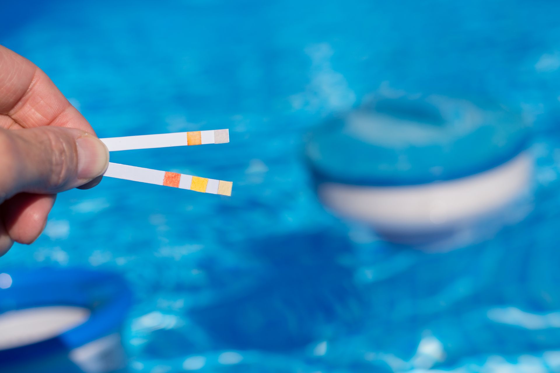 Hand haelt zwei Teststreifen zur Messung von pH-Wert und Chlorgehalt vor klarem Poolwasser
