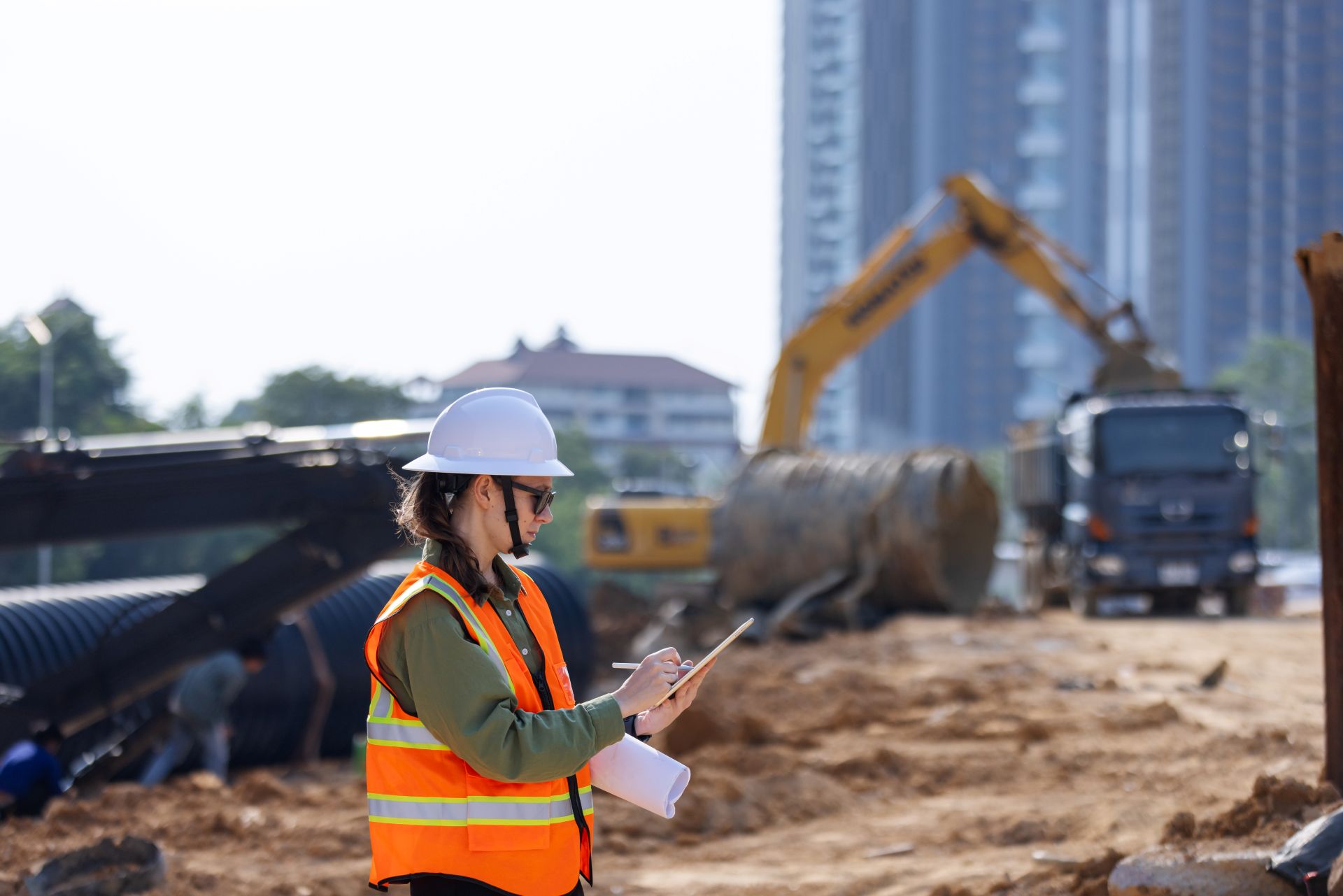 Bauleiterin mit Schutzhelm und Tablet auf Baustelle eines unterirdischen Entwässerungssystems mit Rohren und Baggern im Hintergrund